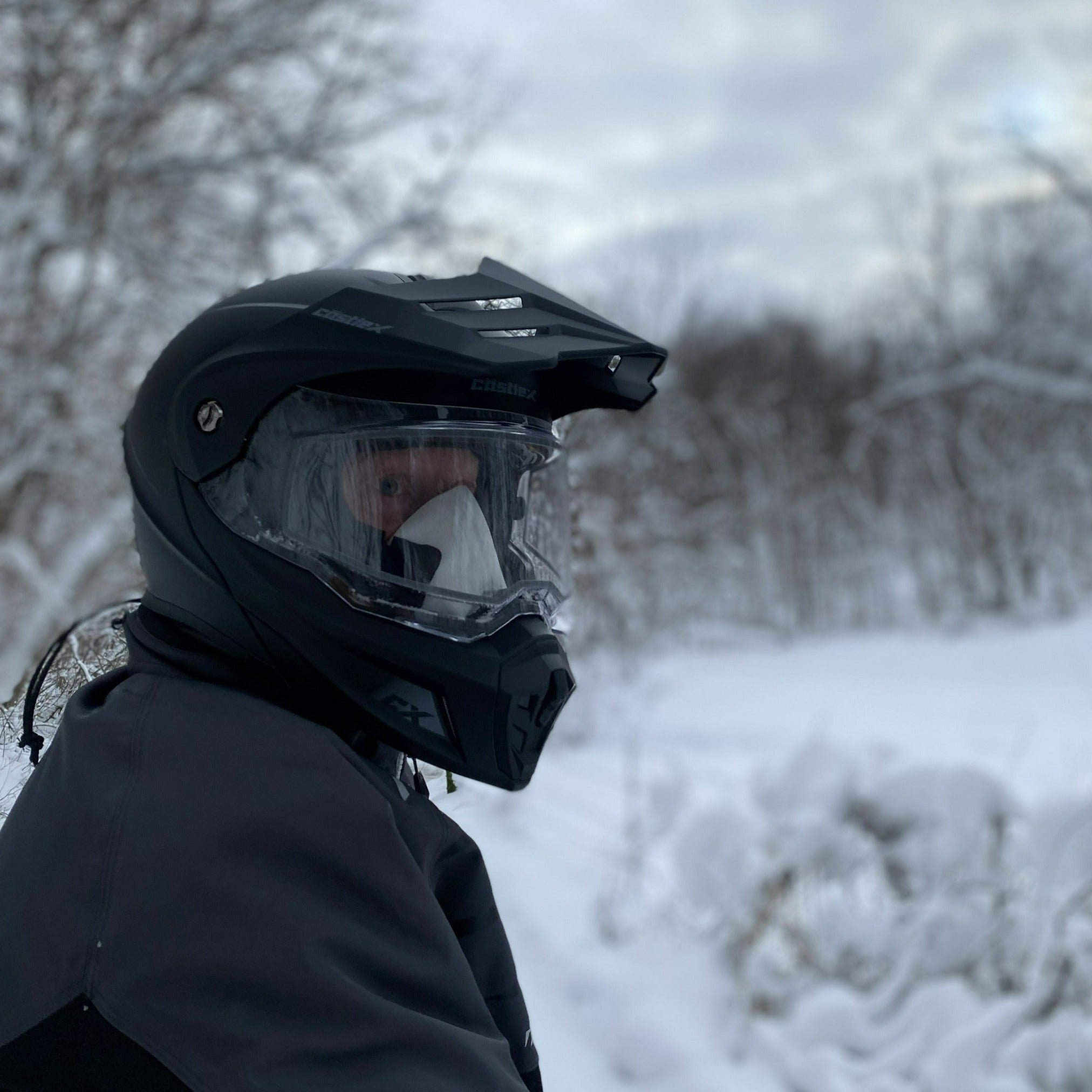 Man wearing a helmet while snowmobiling in a snowy wooded area with a SNOWFLOW balaclava on